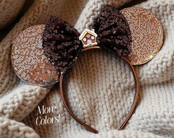 May include: A brown headband with sequined ears and a sequined bow. The ears are gold and brown, and the bow is dark brown. A small gingerbread house charm is in the center of the bow. The text "More Colors!" is visible.
