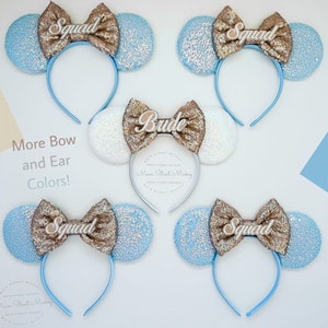 Puede incluir: Cuatro pares de orejas de Minnie Mouse de lentejuelas azul claro y doradas con una diadema. Las orejas están decoradas con un lazo dorado y un letrero de madera blanco con las palabras "Squad" o "Bride".