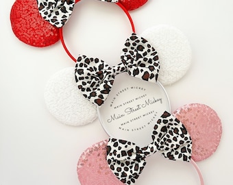 May include: Two pairs of Minnie Mouse ears with leopard print bows. The ears are red, white, and pink with sequins. The headband is pink.
