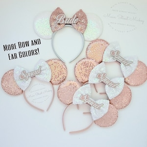 Puede incluir: Cuatro pares de orejas de Minnie Mouse de lentejuelas rosas y blancas con una diadema. Las orejas están decoradas con un lazo blanco y una palabra de purpurina dorada. Las palabras son "Bride", "Squad", "Squad" y "Squad".