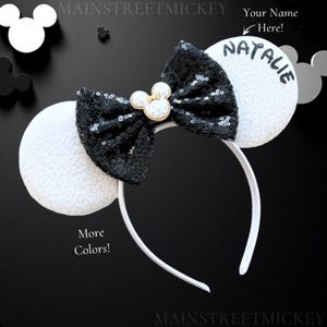 Puede incluir: Diadema de orejas de Minnie Mouse blancas con un lazo de lentejuelas negras y un dije de Mickey Mouse de perla. Las orejas están personalizadas con el nombre "Natalie".