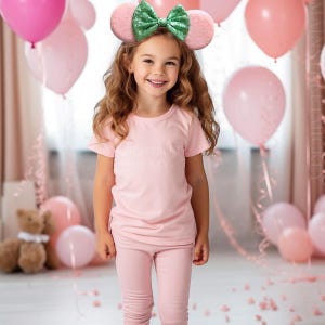 May include: A young girl wearing a pink outfit and a headband with mouse ears and a green sequined bow. The pink outfit includes a t-shirt with the text "MAIN STREET MICKEY" and matching leggings. Pink balloons are in the background.