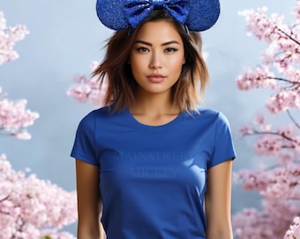 May include: A woman wearing a blue t-shirt with the text "Main Street Mickey" printed on the front. She is wearing blue glitter Minnie Mouse ears with a large bow.