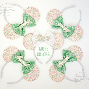Puede incluir: Cuatro pares de orejas de Minnie Mouse en verde pastel y blanco con un lazo verde y letras doradas. Las letras dicen "Bride" y "Squad".