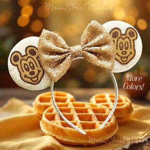 May include: Gold sequin Minnie Mouse ears with a brown Mickey Mouse silhouette on each ear. The ears are on a white headband and have a large gold bow.