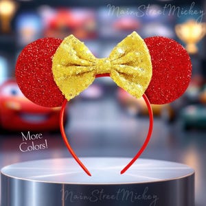May include: Red Minnie Mouse ears headband with a large gold sequin bow. The headband is on a silver display stand. The text "Main Street Mickey" is visible in the background.