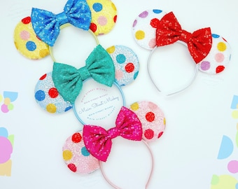 May include: Three pairs of colorful polka dot Minnie Mouse ears with bows. The ears are on white headbands. The bows are blue, teal, and pink.