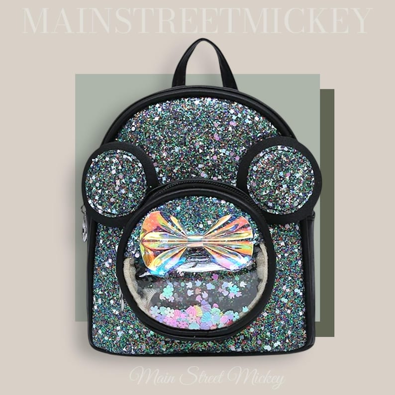 May include: A black glitter backpack with a clear plastic window on the front. The window shows a rainbow bow and colourful confetti. The backpack has black ears on the top and the text "Main Street Mickey" is printed on the bottom.