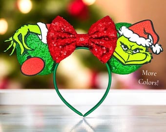 May include: Green and red sequined Minnie Mouse ears with a Grinch character wearing a Santa hat on one ear. The bow is red and the headband is green. The text "More Colors!" is visible in the image.