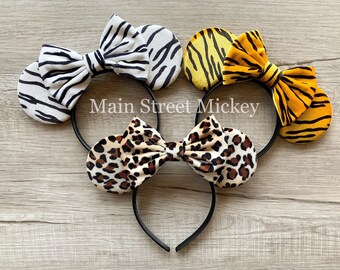 May include: Three pairs of fabric-covered mouse ears with bows. The ears are zebra print, leopard print, and tiger print. The bows are the same print as the ears. The ears are attached to a black headband. Main Street Mickey is written on the headband.