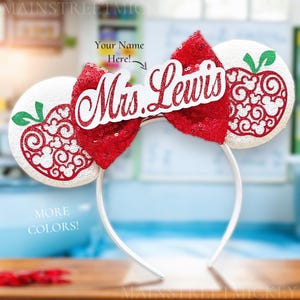 May include: White Minnie Mouse ears with a red sequined bow and a white apple cutout with red swirls. The ears have a white name tag that says "Mrs. Lewis".