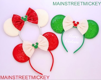May include: Two pairs of festive Minnie Mouse ears with red, white, and green sequin bows. The ears are on a white background.