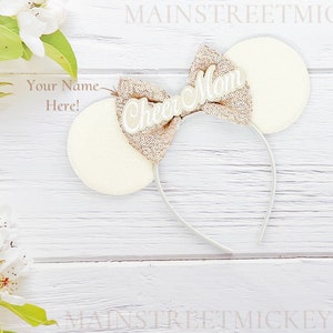 May include: Cream-colored mouse ear headband with a sequined bow that reads "Cheer Mom" in white script. The ears are round and the headband is white. The background is a white wooden surface with floral accents.