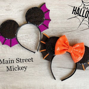 May include: Two pairs of black and purple Mickey Mouse ears with bat wings. One pair has a black and orange bow. The text "Main Street Mickey" is written on a wooden surface. The text "Happy Halloween" is written on a white background with a black spider web.