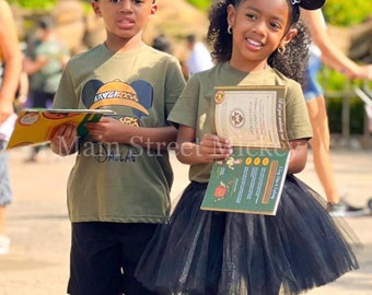 May include: Two children in olive green t-shirts with a cartoon design, black shorts, and white trainers. They wear Mickey Mouse ear headbands. One child has a black tutu. The text "Main Street Mickey" is visible.