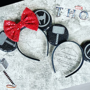 Pode incluir: Orelhas de Minnie Mouse pretas e prateadas com um laço vermelho. As orelhas apresentam os personagens da Marvel Thor e Loki. A faixa de cabeça é preta e diz "Main Street Mickey" em branco.