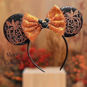 May include: Black headband with sequined ears and an orange sequined bow. The ears feature a pumpkin design. A black Mickey Mouse emblem is centered on the bow. The text "More Colors!" is visible.