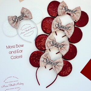 Puede incluir: Cuatro orejas de Minnie Mouse de lentejuelas rojas y doradas con texto negro que dice "Squad" en cada diadema. Una diadema de orejas de Minnie Mouse de lentejuelas blancas con texto negro que dice "Bride".
