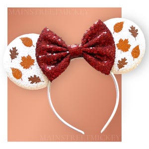 May include: White Minnie Mouse ears with a red sequined bow and a fall leaf design. The leaves are in shades of brown and orange.