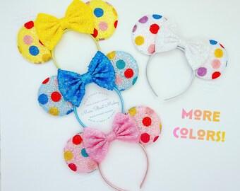 May include: Three pairs of colorful polka dot Minnie Mouse ears with bows. The ears are made of sequins and have a headband. The colors are yellow, blue, and pink.