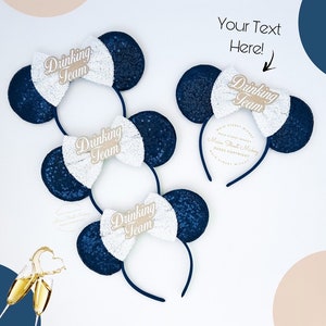 May include: Set of three blue and white sequined mouse ear headbands with a gold bow and the text "Drinking Team" on each headband.