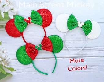 May include: Two pairs of Minnie Mouse ears with a bow. One pair is red and green with gold accents. The other pair is white with green and gold accents. The text "More Colors!" is written on the white wooden background.