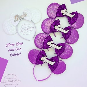 Puede incluir: Cuatro orejas de Minnie Mouse de lentejuelas moradas con lazos blancos y la palabra "Squad" en cada lazo. Una diadema de orejas de Minnie Mouse de lentejuelas blancas con un lazo blanco y la palabra "Bride" en el lazo. El texto "Main Street Mickey" está en la diadema.