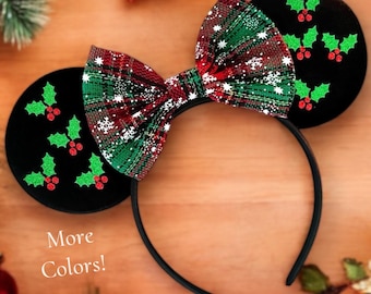May include: Red and green plaid bow with white snowflakes and holly leaf embellishments on a black headband. The headband is decorated with green holly leaves and red berries. The text "More Colors!" is visible in the image.