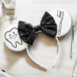 May include: White mouse ears with a black bow. One ear has a cartoon tooth with a smiling face. The other ear has a cartoon toothbrush. The ears are attached to a white headband. "More Colors!" is written on the ear with the tooth.