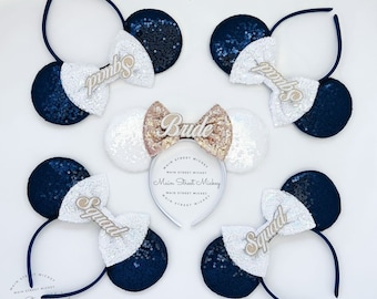 May include: Set of five navy blue and white sequined Minnie Mouse ears headbands with gold glitter lettering. One headband says "Bride" and the other four say "Squad".