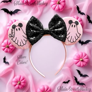 May include: A Halloween-themed headband with a black sequined bow and pink ears featuring a ghost design and the word "Boo". The headband is surrounded by pink pumpkins and black bat cutouts. The text "More Colors!" is also visible.