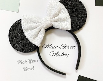 May include: Black sequined mouse ears with a white sequined bow on a black headband. The text "Main Street Mickey" and "Pick Your Bow!" are visible on the white background.