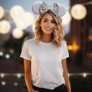 Puede incluir: Una mujer lleva una camiseta blanca con el texto "Mainstreet Mickey" impreso en ella. Lleva una diadema plateada y blanca con un lazo brillante y la palabra "Bride" en ella. La diadema tiene forma de orejas de Mickey Mouse.