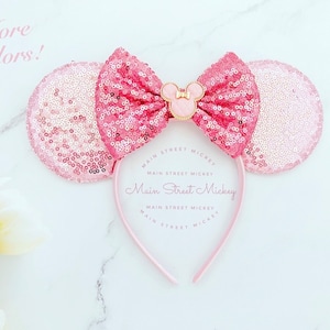 Puede incluir: Diadema de orejas de Minnie Mouse con lentejuelas rosas con un lazo a juego y un pequeño emblema de Minnie Mouse. La diadema es de color rosa claro con el texto "Main Street Mickey". La imagen también incluye el texto "More Colors!"