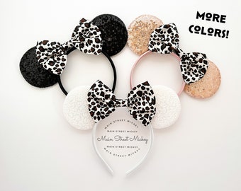May include: Three pairs of Minnie Mouse ears with a leopard print bow and different coloured sequin ears. The ears are black, pink, and white. The headband is clear with the text "Main Street Mickey" printed on it.