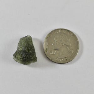 May include: A small, raw, olive-green moldavite stone with a textured, irregular surface positioned next to a silver-coloured United States quarter coin for scale, showing the relative size of the mineral specimen against the 24.26 mm diameter coin.