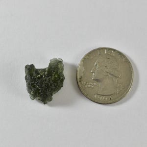 May include: A small, rough, olive-green moldavite stone with a textured, irregular surface next to a silver-coloured United States quarter coin, which has a diameter of approximately 2.43 cm, for scale on a plain white surface.