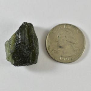 Peut inclure: Une pierre de moldavite brute vert olive foncé avec une surface texturée et irrégulière à côté d'une pièce de monnaie américaine de 25 cents pour l'échelle, montrant que la pierre mesure un peu moins de 2,43 cm de diamètre.