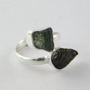 May include: A silver ring with two rough, dark green moldavite stones. The stones are set in bezel settings, and the ring band curves around the finger. The ring is displayed on a white background. The ring is a piece of jewellery.