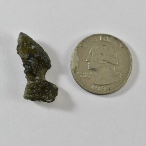 May include: A small, dark olive-green, irregularly shaped piece of moldavite with a textured, pitted surface, positioned next to a silver-coloured United States quarter coin for scale, showing the relative size of the mineral specimen.