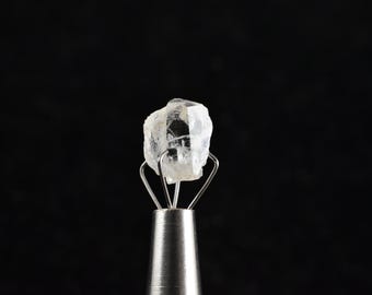 May include: A clear, faceted crystal is held in a metal clamp. The crystal is a light, translucent white.