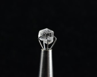 May include: A clear, faceted crystal is held in a metal clamp. The crystal is a hexagonal shape and is translucent. The clamp is silver and has a black background.
