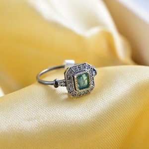 May include: A silver ring with a square emerald green gemstone surrounded by smaller diamonds. The ring is set on a yellow satin fabric.