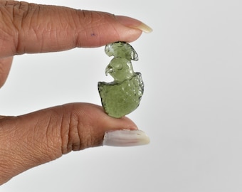 May include: A small, carved green gemstone shaped like a bird hatching from an egg. The gemstone is held between two fingers against a white background. The bird has a detailed beak and head.