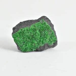 May include: A rough, dark gray rock with a large patch of bright green crystals. The crystals are densely packed and have a sparkly, almost metallic sheen.