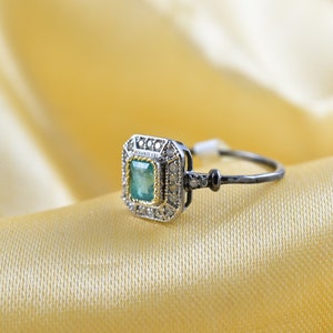 May include: A silver ring with a green emerald gemstone surrounded by smaller diamonds. The ring has a black band.