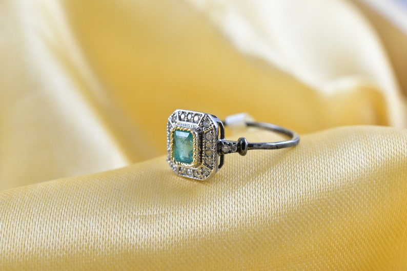 May include: A silver ring with a green emerald gemstone surrounded by smaller diamonds. The ring has a black band.