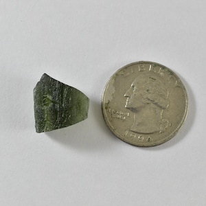 May include: A small, irregular, translucent olive-green stone fragment with a textured, etched surface, positioned next to a silver-coloured United States quarter coin for scale, showing the relative size of the mineral piece.
