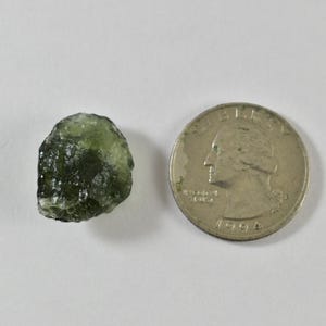 May include: A small, translucent, olive-green raw gemstone with a textured, irregular surface next to a silver-coloured United States quarter coin for scale. The stone displays varying shades of deep green and has a glassy, pitted appearance.