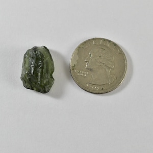 May include: A small, raw, translucent olive-green moldavite stone with an irregular, textured surface. The stone is positioned next to a silver-coloured United States quarter coin, which serves as a visual reference for the stone's size.